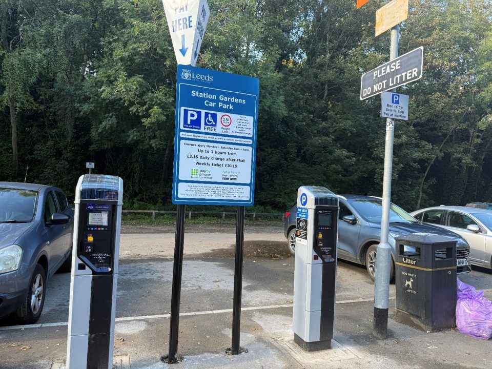 A car park scene at Station Gardens Car Park in Leeds, UK, featuring two modern pay-and-display parking meters in the foreground. A prominent blue sign provides parking information, stating that parking is free for up to 3 hours from Monday to Saturday (8am–6pm), with a £2.15 daily charge thereafter and an optional £10.15 weekly ticket. A small sign above the meters reads "PLEASE DO NOT LITTER" and another one details the pay times. Nearby are parked vehicles and a black litter bin marked for dog waste. The area is bordered by dense green trees in the background.