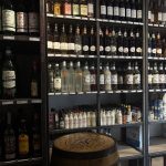 The image shows a well-organized display of alcoholic beverages and cocktail ingredients on shelves in a liquor store or specialty shop.