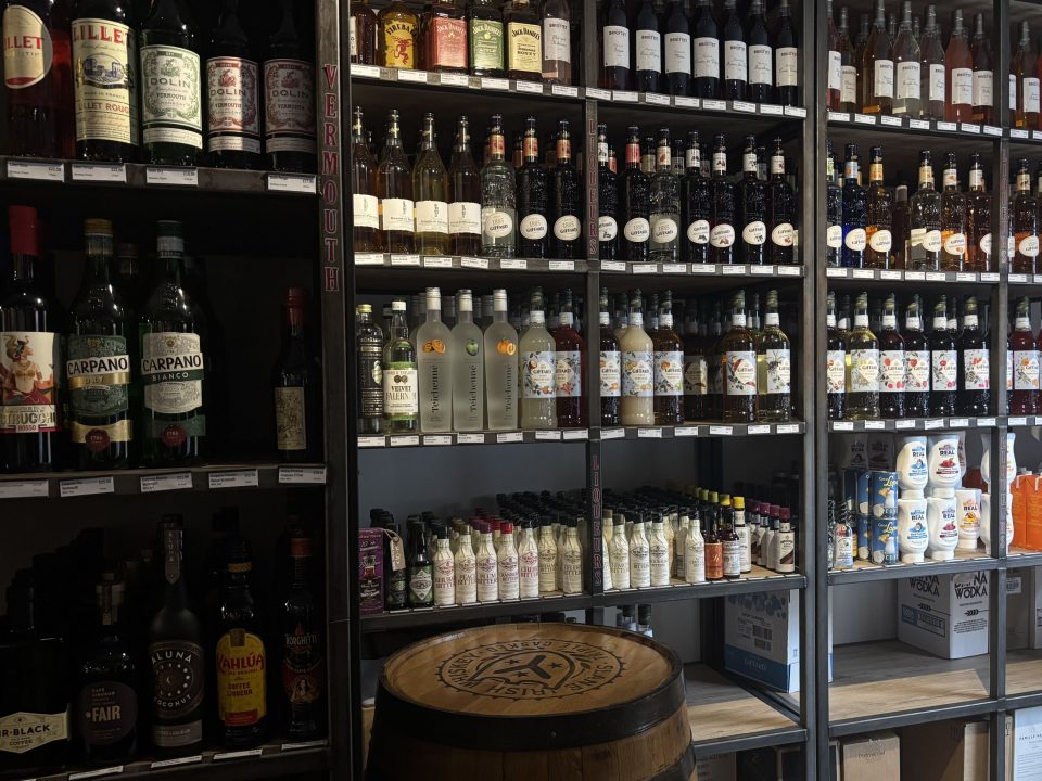 The image shows a well-organized display of alcoholic beverages and cocktail ingredients on shelves in a liquor store or specialty shop.