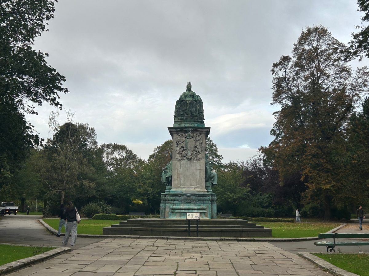image of a statue of queen Victoria in Hyde Park Leeds. there is a stone flag walkway up to the statue and grass and trees surrounding it. Various students are dotted walking around the frame.