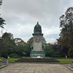 image of a statue of queen Victoria in Hyde Park Leeds. there is a stone flag walkway up to the statue and grass and trees surrounding it. Various students are dotted walking around the frame.