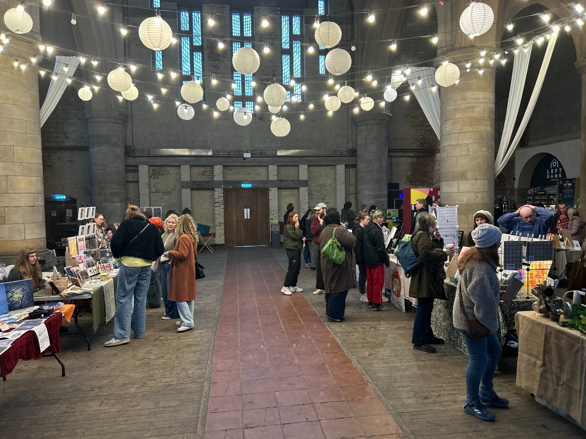 : Indoor market scene inside a large hall with tall stone columns and hanging string lights with paper lanterns. People browse various craft and art stalls displaying prints, handmade goods, and other items. The atmosphere is warm and lively, with vendors interacting with visitors under soft lighting.