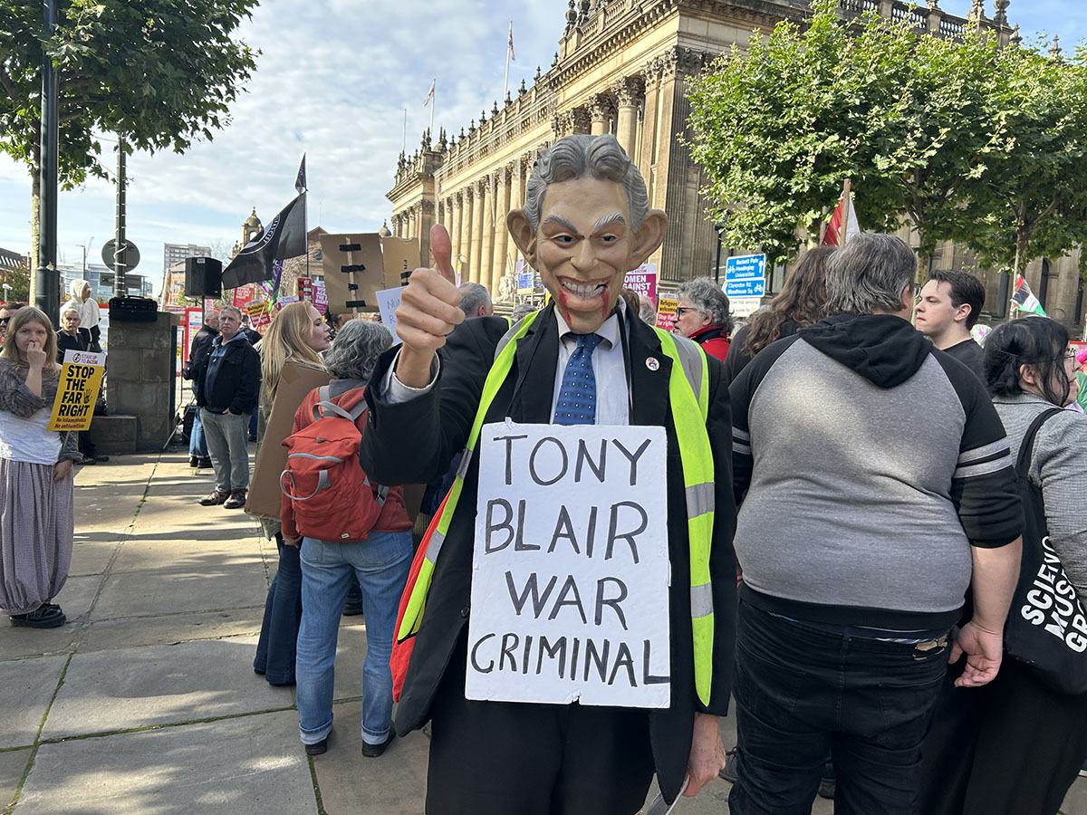 Man wearing suit and Tony Blair mask with fake blood coming out of mouth. Sign around neck saying "Tony Blair war criminal"