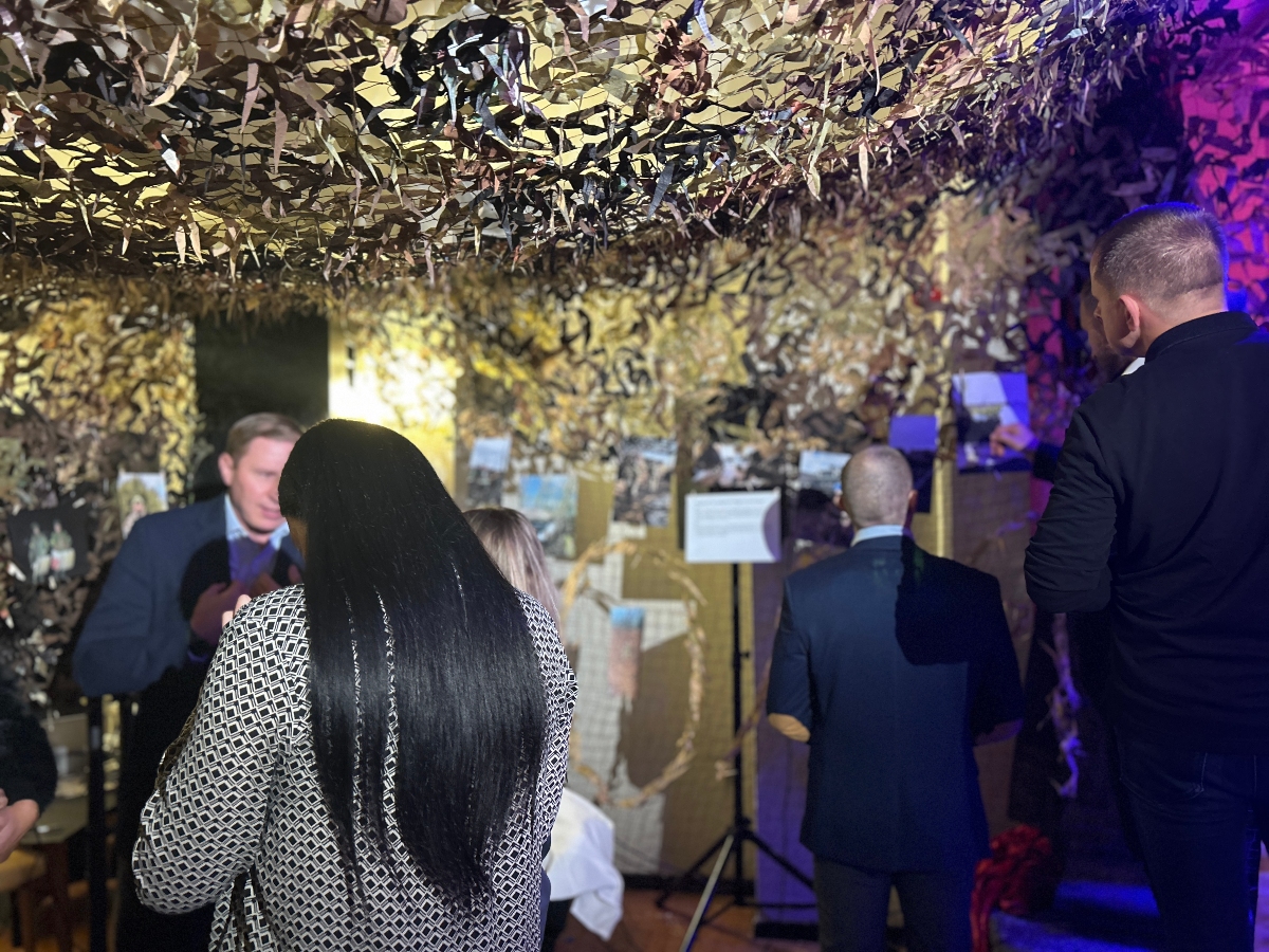 A group of people view a war-themed exhibit covered in camouflage netting on the ceiling and walls. Photographs and informational posters are displayed throughout the space. The lighting is dim with blue and yellow tones creating a dramatic atmosphere. The visitors, dressed in formal and semi-formal attire, appear engaged as they examine the displays.