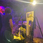 Two people, a man wearing a ceremonial chain and a woman in a black dress, stand closely together viewing a wartime-themed display. The dimly lit exhibit features camouflage netting, photographs, and a headless mannequin dressed in a suit with a Ukrainian flag draped over the shoulders. A tripod stands nearby, and the scene is illuminated with dramatic purple and yellow lighting.