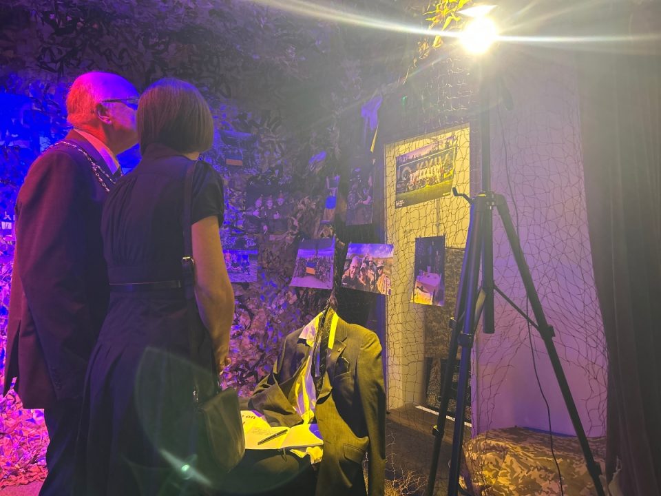 Two people, a man wearing a ceremonial chain and a woman in a black dress, stand closely together viewing a wartime-themed display. The dimly lit exhibit features camouflage netting, photographs, and a headless mannequin dressed in a suit with a Ukrainian flag draped over the shoulders. A tripod stands nearby, and the scene is illuminated with dramatic purple and yellow lighting.