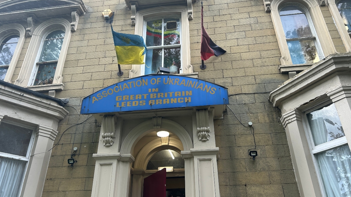 Front entrance of the Association of Ukrainians in Great Britain, Leeds Branch. The building is a traditional stone structure with arched windows. Above the doorway is a blue sign with yellow text. Two flags are displayed above the entrance: the blue and yellow Ukrainian national flag and a red-and-black flag representing Ukrainian nationalism. A window above the sign has a rainbow flag visible behind the glass.