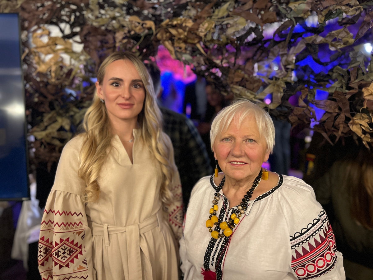 Olga Callaghan and Dariia Kovnatska. Two individuals wearing traditional embroidered clothing stand in front of a decorative backdrop made of camouflage netting and foliage. The person on the left wears a beige outfit with red and white embroidery, while the person on the right dons a white garment with black and red embroidery and a necklace of large yellow beads. The setting appears festive, with colorful lighting suggesting a cultural or celebratory event."