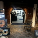 A gallery with brick walls and arched doorways showcasing large, detailed photographs of human irises, with a sign reading "VIZUALYEYES" and prices starting from £29.