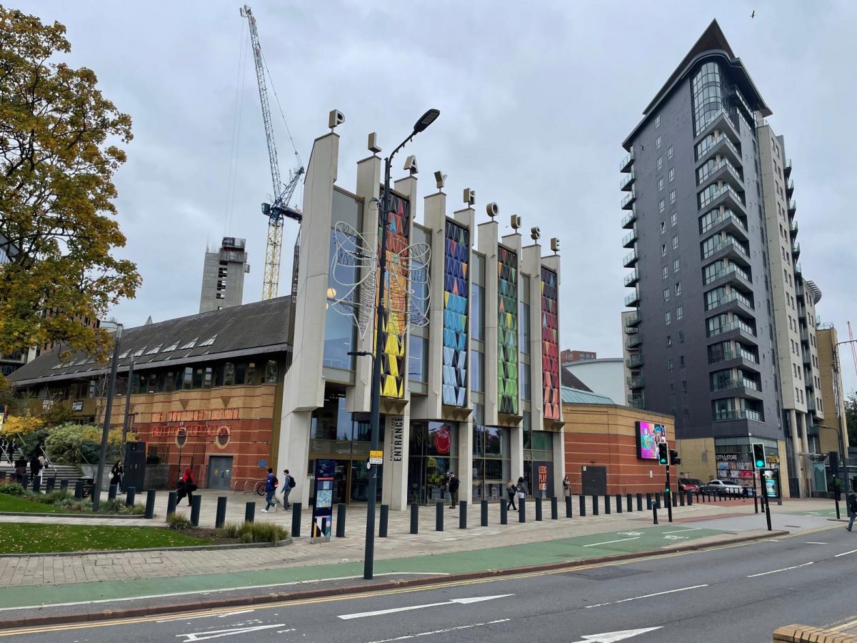 The image shows a city street view featuring a modern building with tall, colorful panels on its facade and a sign reading “Front Room Café Entrance.” The structure is part of a larger complex with “The Quarry Theatre” signage visible on the adjoining section. Pedestrians walk along the pavement, and a tall residential tower stands beside the building. A construction crane and other buildings are visible in the background under an overcast sky.