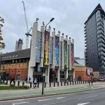 The image shows a city street view featuring a modern building with tall, colorful panels on its facade and a sign reading “Front Room Café Entrance.” The structure is part of a larger complex with “The Quarry Theatre” signage visible on the adjoining section. Pedestrians walk along the pavement, and a tall residential tower stands beside the building. A construction crane and other buildings are visible in the background under an overcast sky.