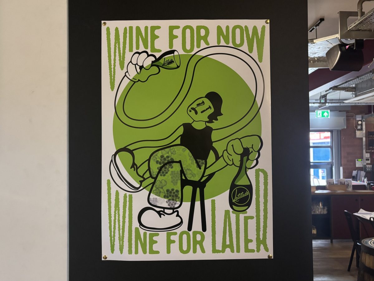 The image shows a colorful, stylized poster mounted on a dark wall inside a shop or bar. The poster features a cartoon-style illustration of a relaxed character sitting on a chair, holding a wine bottle labeled “Latitude” in one hand and pouring wine into a glass with the other. The background is a large green circle, and the text on the poster reads: “WINE FOR NOW, WINE FOR LATER.”