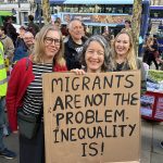 Three women holding cardboard sign saying "migrants are not the problem. inequality is!" in capital letters