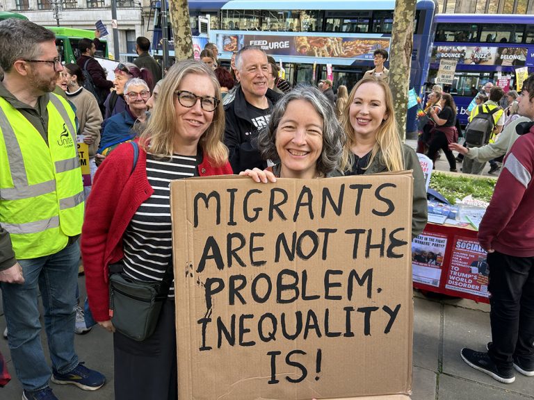 Three women holding cardboard sign saying "migrants are not the problem. inequality is!" in capital letters