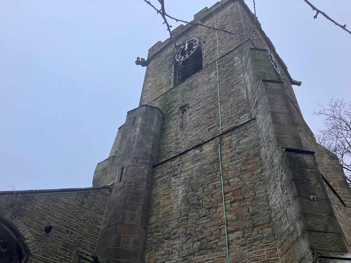 A close-up upward view of a stone church tower with a clock face near the top. The structure has weathered stone walls, narrow arched windows, and a battlemented roofline. The image captures the height and texture of the tower against an overcast sky, with some branches visible at the top edge.