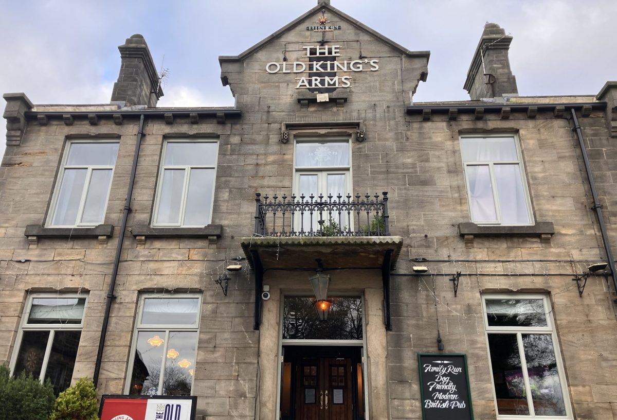 A large two-story stone pub building with tall rectangular windows and a central entrance beneath a small balcony. The sign at the top reads “THE OLD KING’S ARMS.” A hanging lantern is above the doorway, and string lights are draped across the facade. To the left of the entrance is a signboard with the pub’s logo, and to the right, a chalkboard-style sign advertises that it is a family-run, dog-friendly British pub. Some greenery is in front of the building, and a person with a camera is visible in the lower left corner.