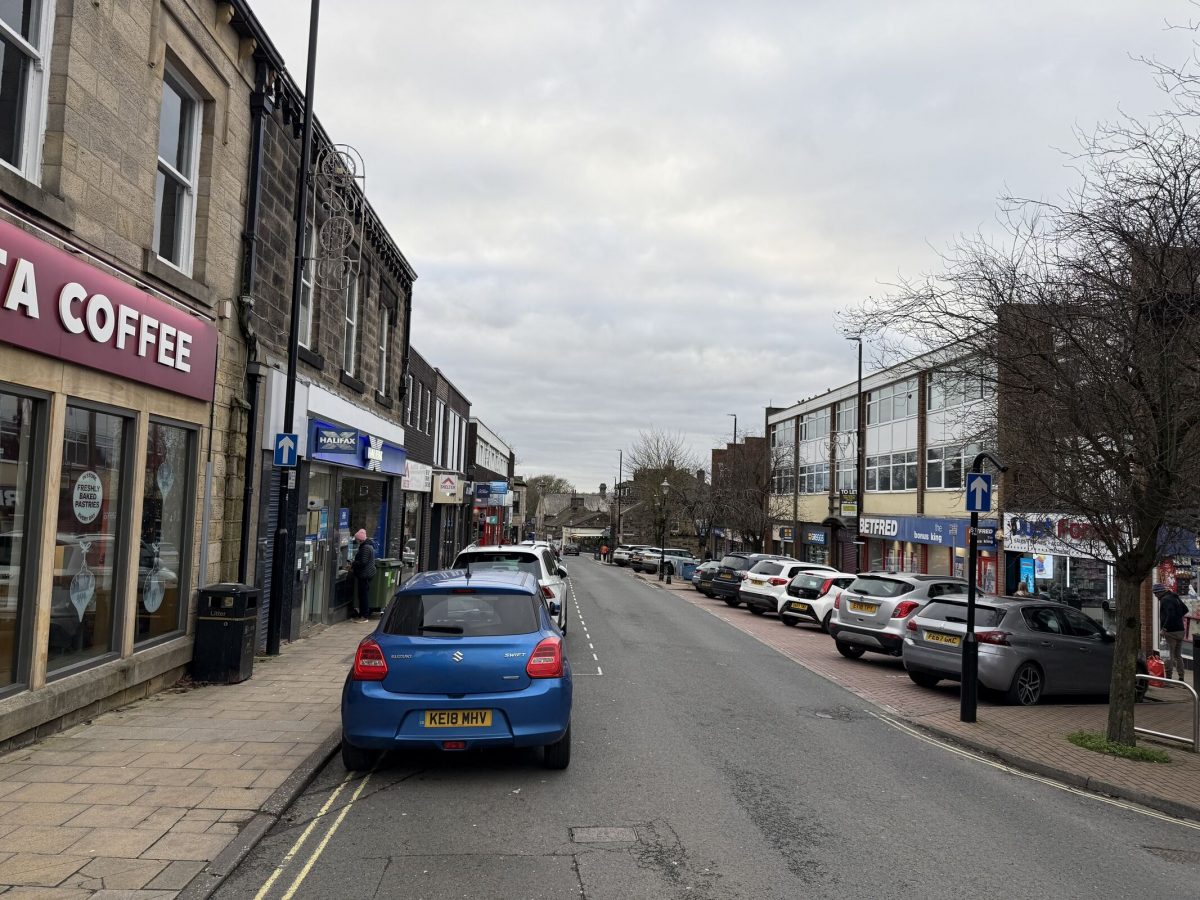 A small town street with parked cars lining both sides. On the left are shops including a Costa Coffee and a Halifax bank ATM where a person is using the machine. On the right are more shops such as Betfred and a convenience store. The sky is overcast, and a few pedestrians are walking along the pavements. The street is one-way, indicated by blue arrow signs, and the buildings have a mix of stone and brick façades.
