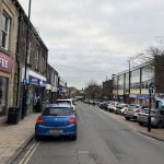 A small town street with parked cars lining both sides. On the left are shops including a Costa Coffee and a Halifax bank ATM where a person is using the machine. On the right are more shops such as Betfred and a convenience store. The sky is overcast, and a few pedestrians are walking along the pavements. The street is one-way, indicated by blue arrow signs, and the buildings have a mix of stone and brick façades.