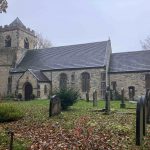 A stone church building with a clock tower, surrounded by a graveyard. The church has a traditional Gothic design with arched windows. Autumn leaves cover the ground, and the scene appears overcast, creating a serene and slightly somber atmosphere.