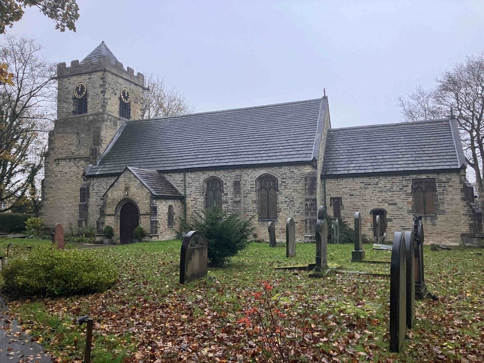 A stone church building with a clock tower, surrounded by a graveyard. The church has a traditional Gothic design with arched windows. Autumn leaves cover the ground, and the scene appears overcast, creating a serene and slightly somber atmosphere.