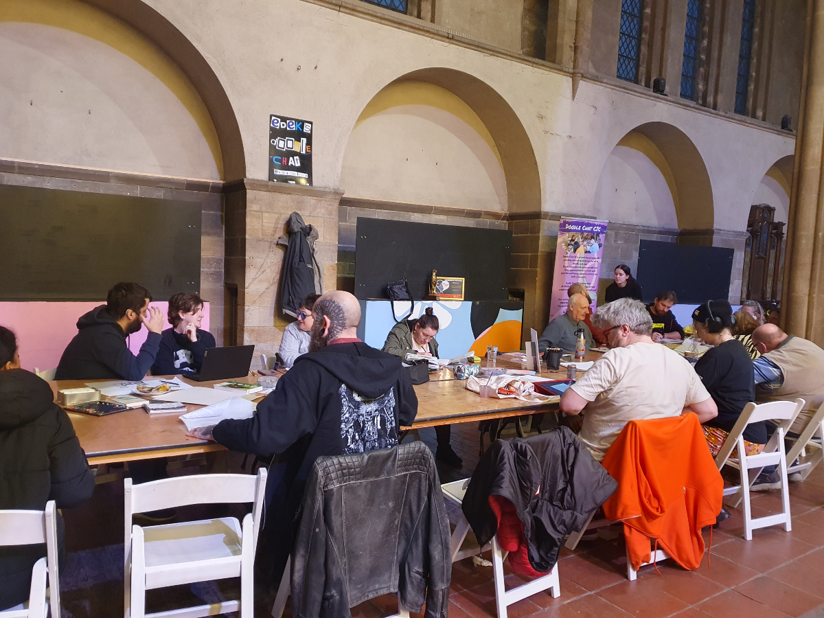 The image shows a group of people drawing around a table with jackets hanging over the back of chairs