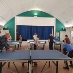 The image shows players in a church hall playing table tennis
