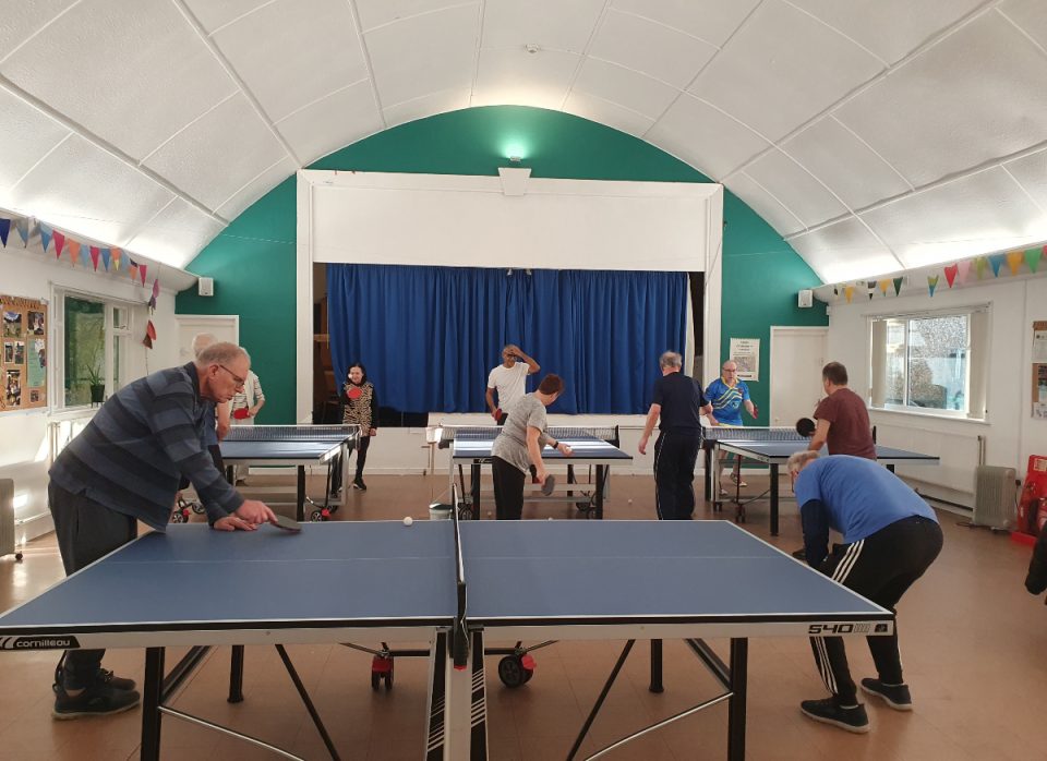 The image shows players in a church hall playing table tennis