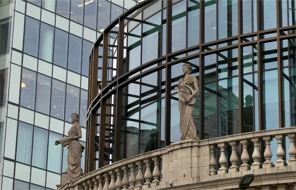 Stone statues of two robed female figures stand on the balustrade of a curved building façade, with a modern glass-and-steel office tower rising behind them.