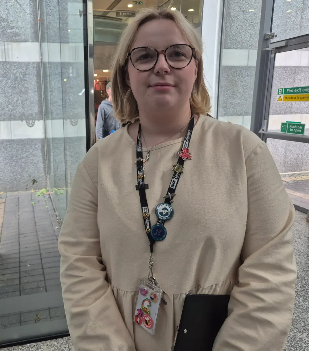 Molly Fielding. Blond hair, glasses, beige shirt; wearing a landyard and holding a clipboard.