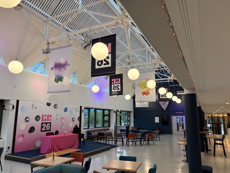 Inside of a building, pink background on display with tables and chairs in the foreground. flags hanging from the ceiling with text on it.