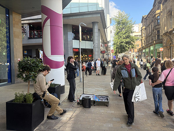 A busy pedestrian shopping street in a city centre. In the foreground, a person carrying several shopping bags walks past a modern building with a large pink-and-white column. To the left, another person sits on a planter looking at a phone, while a street performer stands nearby with a microphone stand, speaker, and equipment case. Numerous shoppers and pedestrians move along the paved walkway between contemporary retail buildings and older stone-fronted architecture in bright daylight.