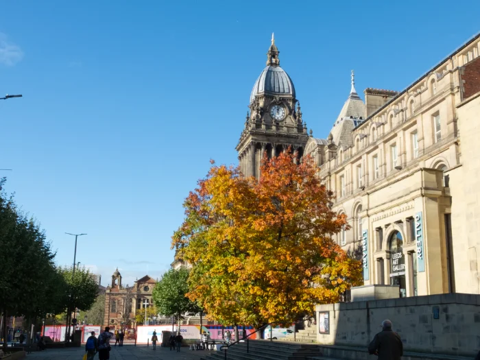 Exterior street scene featuring a tree with vibrant autumn leaves in front of the Leeds Town Hall clock tower and Leeds Art Gallery under a clear blue sky.