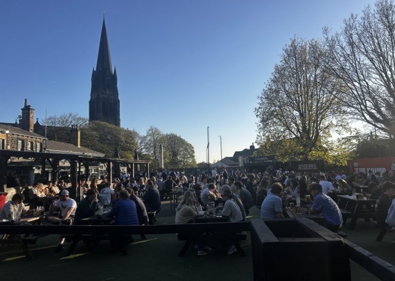 A crowded, sunny outdoor pub beer garden is filled with people sitting at numerous wooden picnic tables on a green artificial turf lawn