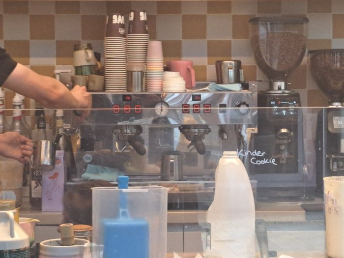 A café counter with a stainless-steel coffee machine, coffee grinders, stacked takeaway cups and milk bottles. A barista prepares a drink behind the counter, with a tiled wall in the background.