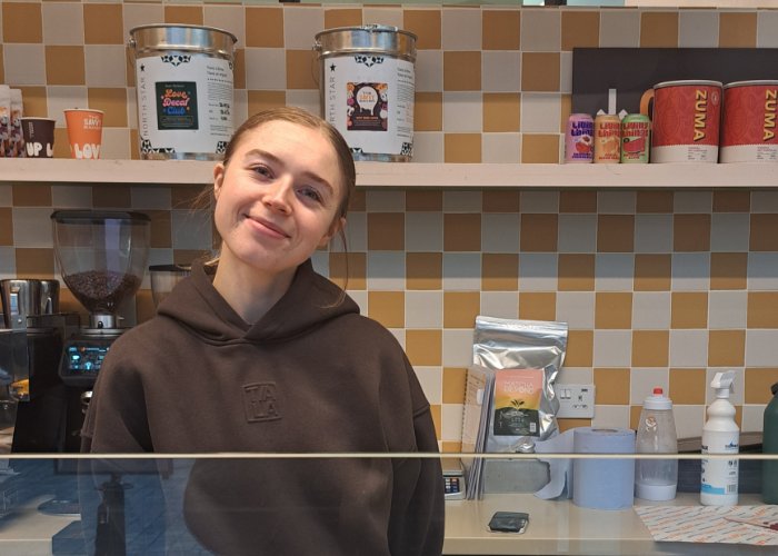 A smiling staff member standing behind the counter at The Savvy Baker, with coffee equipment baking ingredients, with a white and yellow tiled wall behind her.
