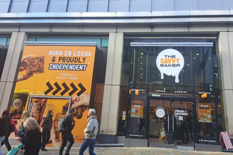 the Savvy Baker storefront, with an orange display window with the words "Born in Leeds and proudly independent". there the white store logo above the glass doors.