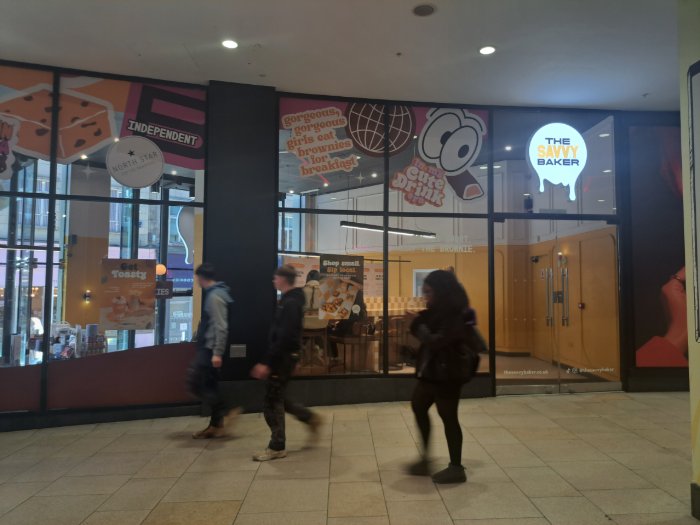 A storefront of The Savvy Baker in an indoor shopping arcade, with orange and pink dessert-themed window graphics, and three people walking past.
