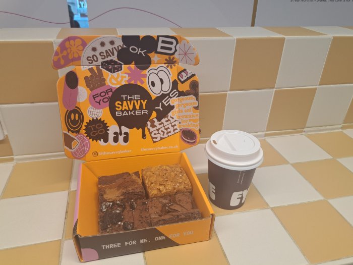 An orange Savvy Baker box containing four assorted brownies sits beside a takeaway coffee cup on a white-and-yellow tiled counter. The box lid features colourful, playful branding.